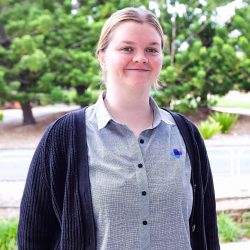 Receptionist headshot, wearing grey shirt with navy blue cardigan, outside with trees in the background.